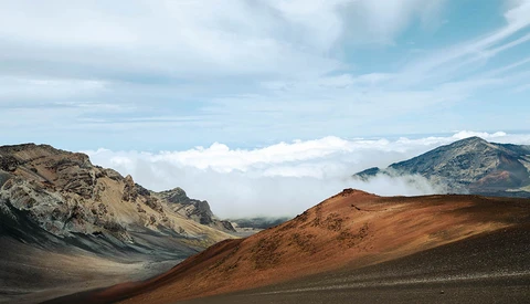 Volcanic landscape with layered rust and charcoal-colored cinder cones beneath a cloud-filled sky.
