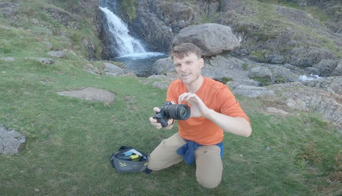 Photographer kneeling outdoors holding a camera, with a small blue camera bag beside him and a waterfall visible in the background.