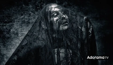 Figure draped in tattered black fabric against dark textured background with dramatic side lighting.