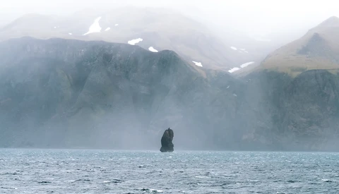 Solitary sea stack rising from misty water with snow-capped mountains shrouded in fog in the background.