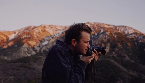 Photographer holding a telephoto lens to his eye, positioned against a mountainous desert landscape at golden hour.