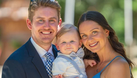 Smiling family of three poses together outdoors in natural sunlight.