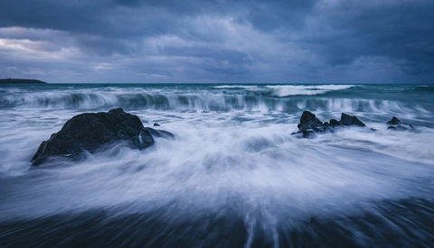 Waves crash over dark rocks on a beach under a dramatic stormy sky.