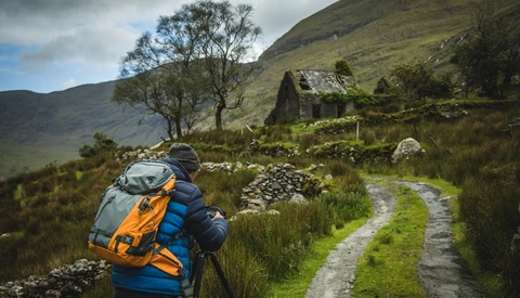 Backpacker hiking through moorland valley with overgrown stone ruins and grassy path.