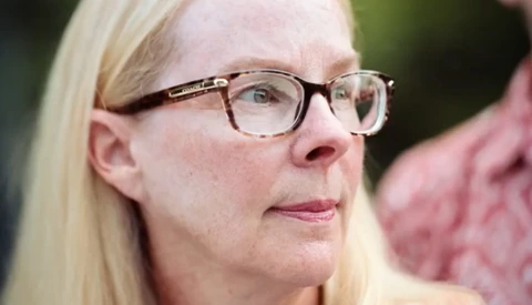 Woman wearing eyeglasses with blonde hair, looking thoughtfully to the side against a blurred green background.