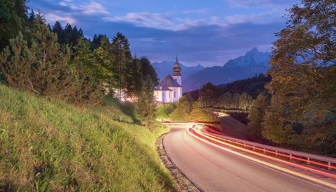 Illuminated church with spire surrounded by forested hills and a winding road with light trails at dusk.