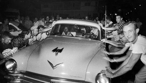 Large crowd surrounds a vintage sedan at night, with people leaning toward and touching the car's exterior.