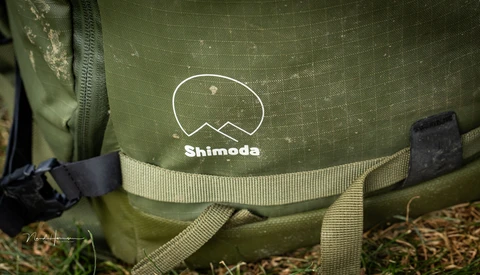 Close-up of a green Shimoda camera bag showing the brand logo and webbing straps with black plastic hardware.