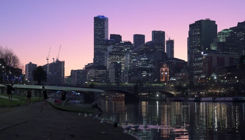 City skyline at twilight with illuminated skyscrapers reflected in still water below a bridge.