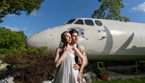 Couple posing in front of a large white aircraft fuselage in a garden setting.