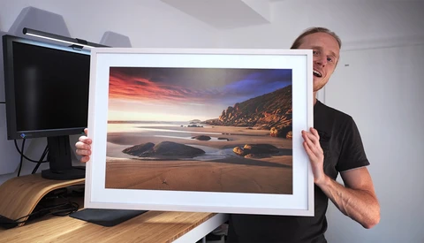 Man holding framed landscape photograph of rocky beach at sunset with dramatic orange and blue sky.