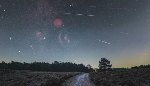 Meteor shower streaking across a twilight sky above a rural landscape with a stream and silhouetted trees.