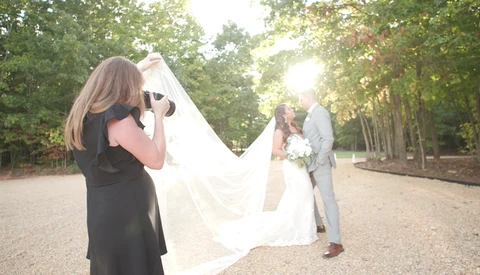 Wedding photographer capturing bride and groom on tree-lined path during golden hour.