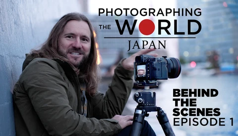 Photographer with long hair holding a medium format camera on tripod, smiling at camera in urban setting.