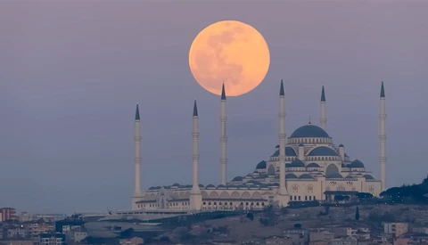 Full moon rising behind a mosque with four minarets and a central dome at dusk.