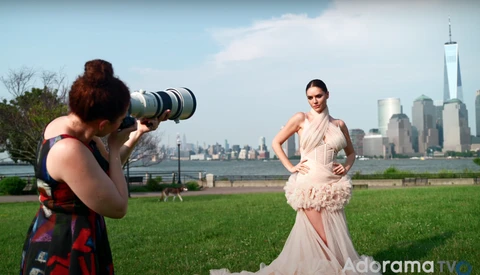 Photographer with telephoto lens capturing portrait of woman in pink gown with NYC skyline backdrop.