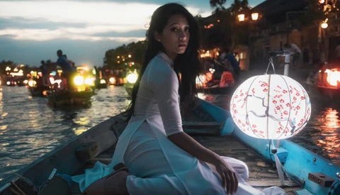 Woman in white clothing holding a decorative lantern in a boat at dusk among illuminated buildings.