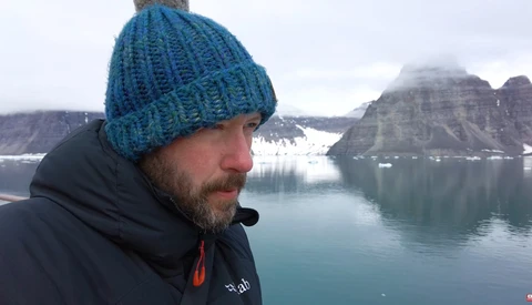 Man in blue knit beanie gazing toward snow-capped mountains across a glacial lake.