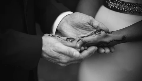 Two people's hands clasped together, one wearing a white sleeve and the other displaying henna designs on dark skin.