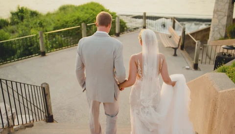 Bride and groom holding hands walking away on an outdoor stone terrace toward a railed overlook.