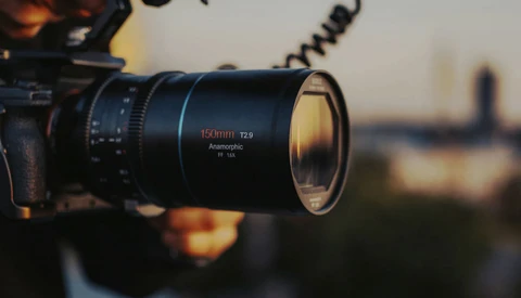 Close-up of a 150mm T2.9 anamorphic lens mounted on a camera, with a blurred cityscape in the background during golden hour.