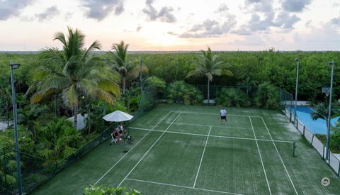 Aerial view of a tennis court with a player mid-game, surrounded by palm trees and lush greenery at dusk.