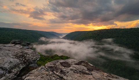 Rocky overlook above a misty river valley at sunrise, with forested hills and dramatic golden-orange clouds.