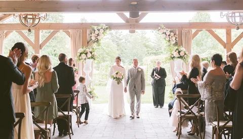 Bride and groom walk down the aisle during a wedding ceremony in a rustic timber-frame venue with guests seated on both sides.