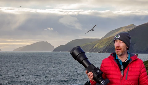 Wildlife photographer holding a telephoto lens while observing a seabird in flight over coastal cliffs.