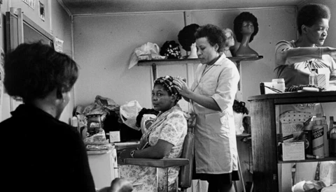 A woman having her hair styled by another woman in what appears to be a beauty salon or barbershop interior.