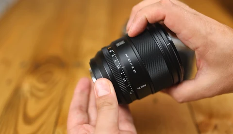 Hand holding a black camera lens with focus ring and depth-of-field markings visible.