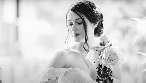 Bride in white lace gown holding bouquet, shot in black and white with soft focus background.