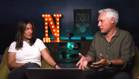 Two people in conversation during an interview or podcast recording set with neon signage in the background.