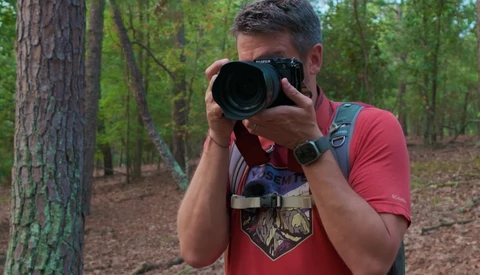 Photographer holding a telephoto lens camera in a forest setting, wearing a red shirt and backpack.