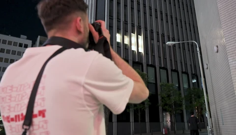 Photographer shooting upward at illuminated modern skyscraper at night with camera raised to eye level.