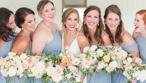 Seven women in matching light blue bridesmaid dresses holding a lush floral arrangement with white, peach, and pink roses.