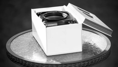 Camera lens in white product box on metallic circular surface against dark background.