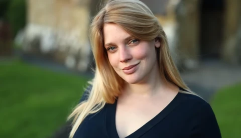 Portrait of a young woman with long blonde hair wearing a black v-neck shirt, photographed outdoors with soft natural light.
