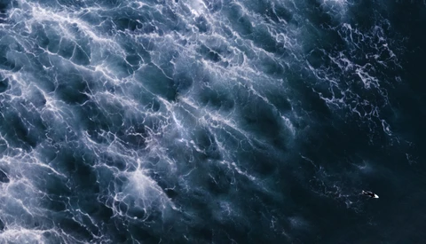 Aerial view of turbulent ocean water with white foam and wave patterns in dark blue-gray tones.