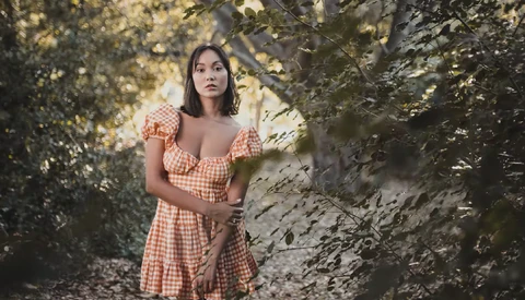 Woman in orange gingham dress posing among green foliage and tree trunks.