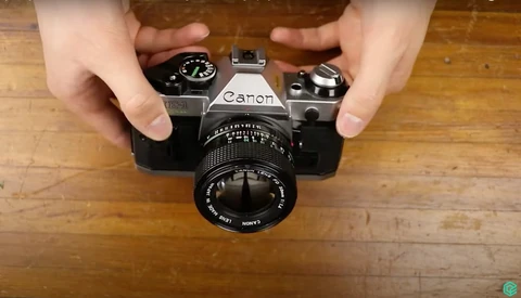Hands holding a vintage Canon SLR camera with manual lens on wooden surface.