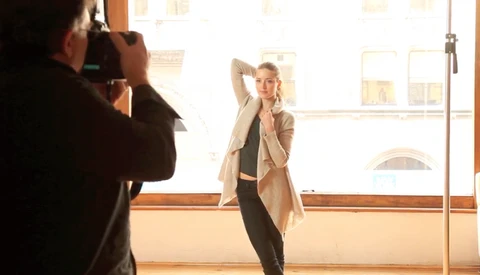 Photographer shooting a portrait of a woman posing in a bright studio with large windows.
