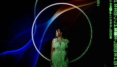 Person in green digital matrix-patterned dress standing within illuminated circular and curved neon light shapes against dark background.