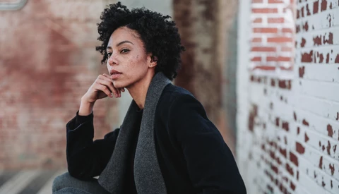 Woman with curly dark hair in thoughtful pose against weathered brick wall.