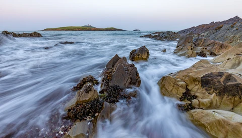 Rocky coastline with flowing water and distant island at twilight.