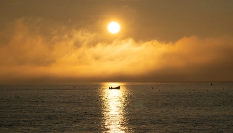 Fishing boat silhouetted against golden sunlight reflecting on calm ocean water at sunset.
