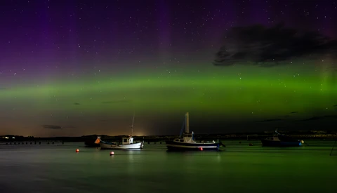 Aurora borealis glowing green and purple above moored boats on calm water at night.