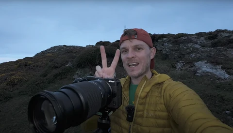 Photographer holding a Nikon camera with telephoto lens on a rocky hillside, making a peace sign at the camera.