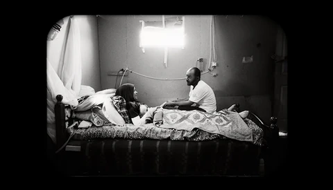 Black and white photograph of two people on a bed in a sparsely furnished room with bright overhead lighting.