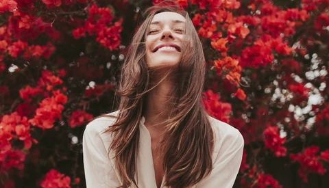 Woman with long brown hair smiling upward among vibrant red flowers in natural sunlight.